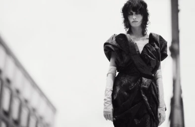 Model in dark sculptural dress with long pale gloves standing against blurred street background