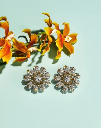 Two flower-shaped gold earrings adorned with clear gemstones sit on a light blue surface, complemented by yellow orchids.