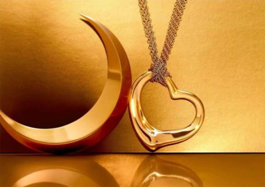 A gold heart pendant necklace hangs near a crescent moon sculpture against a reflective golden background in a jewellery display.
