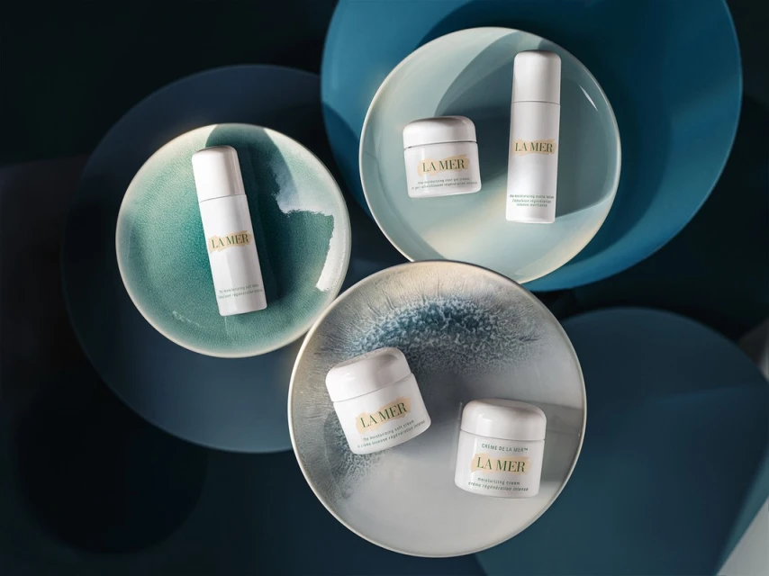 La Mer skincare products are arranged on blue and teal plates, showcasing their moisturizing cream and revitalizing serum in a beauty still life.