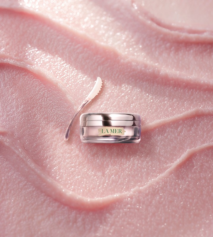 Close-up of La Mer lip balm jar and applicator on light pink background with texture. Focus on beauty and product presentation.