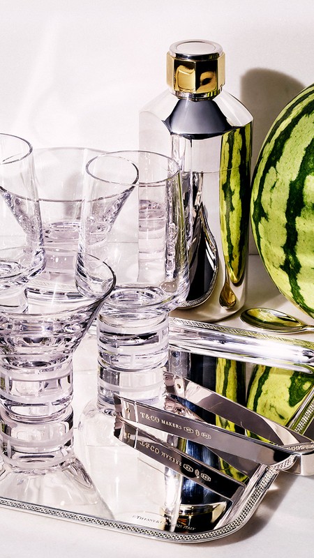 Still life shot of cocktail shaker, cocktail glasses, and watermelon on a silver tray against a white background in a studio.