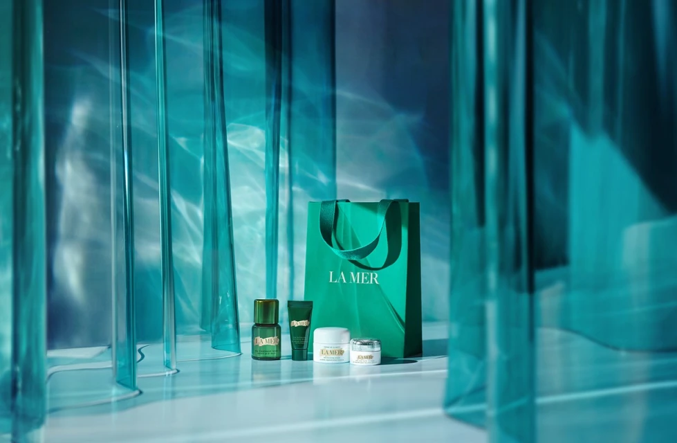 La Mer skincare products and branded shopping bag on a white surface, featuring serum, creams, and lotion, all framed by turquoise rods.