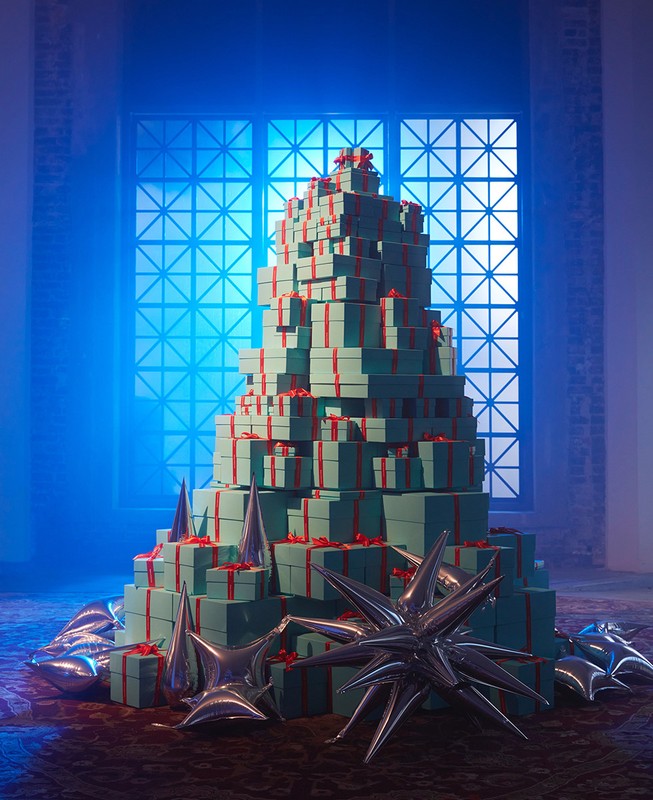 Tower of blue gift boxes with red ribbons, arranged in a tree shape, complemented by star-shaped metallic balloons in an indoor setting.