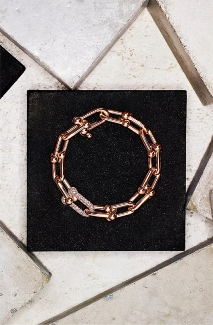 Close-up studio shot of a rose gold chain-link bracelet with diamond-studded clasp on a geometric tile background.
