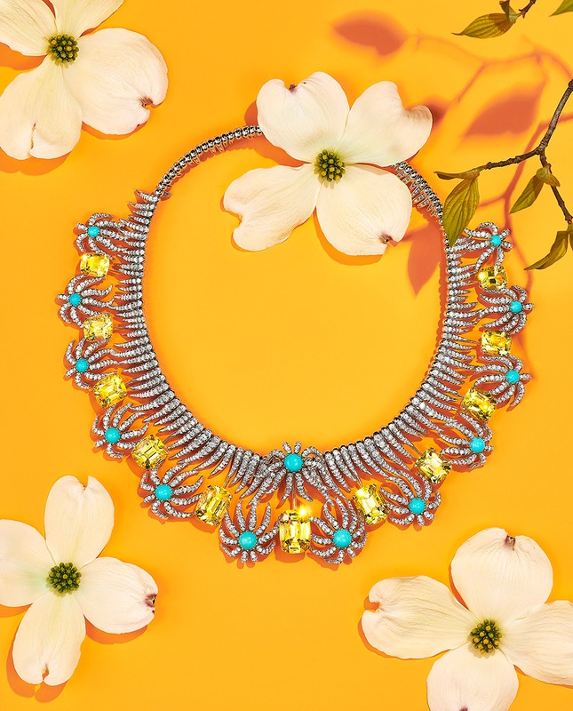 Elegant diamond necklace with turquoise and yellow gemstones surrounded by white dogwood flowers on a bright orange background.