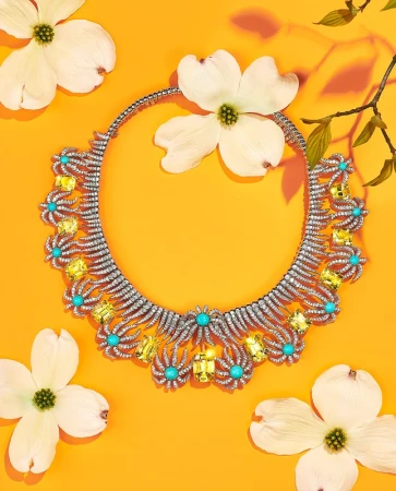 Elegant diamond necklace with turquoise and yellow gemstones surrounded by white dogwood flowers on a bright orange background.