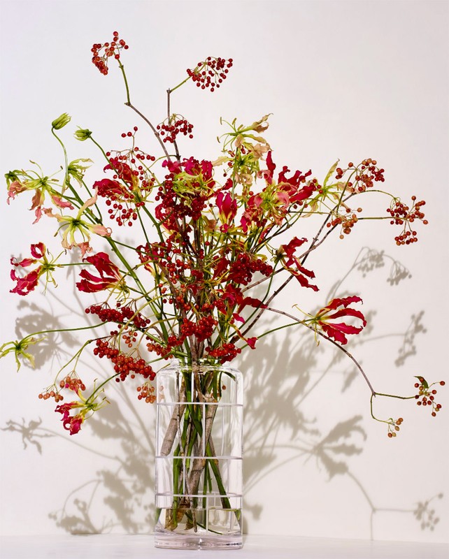 Floral arrangement in a clear glass vase on a white surface with red and green flowers and brown berries against a white backdrop.
