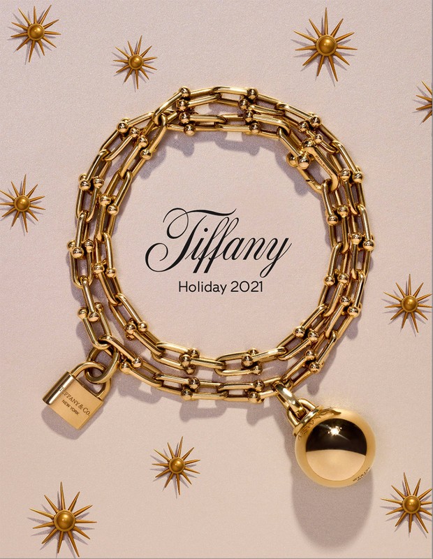 Tiffany & Co. Holiday 2021 campaign featuring a gold chain necklace with padlock and ball pendant on a light pink background with sunbursts.