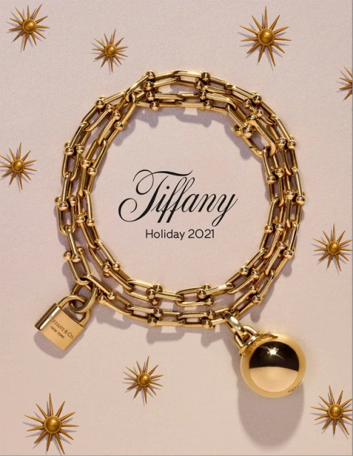 Tiffany & Co. Holiday 2021 campaign featuring a gold chain necklace with padlock and ball pendant on a light pink background with sunbursts.