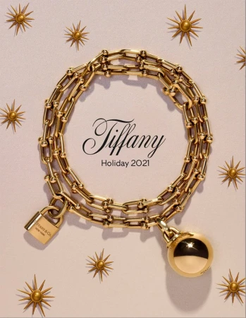 Tiffany & Co. Holiday 2021 campaign featuring a gold chain necklace with padlock and ball pendant on a light pink background with sunbursts.