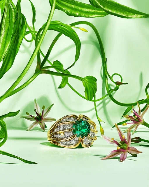 Gold ring with a large green emerald stone surrounded by diamonds, with green leaves and white flowers on a soft green background.