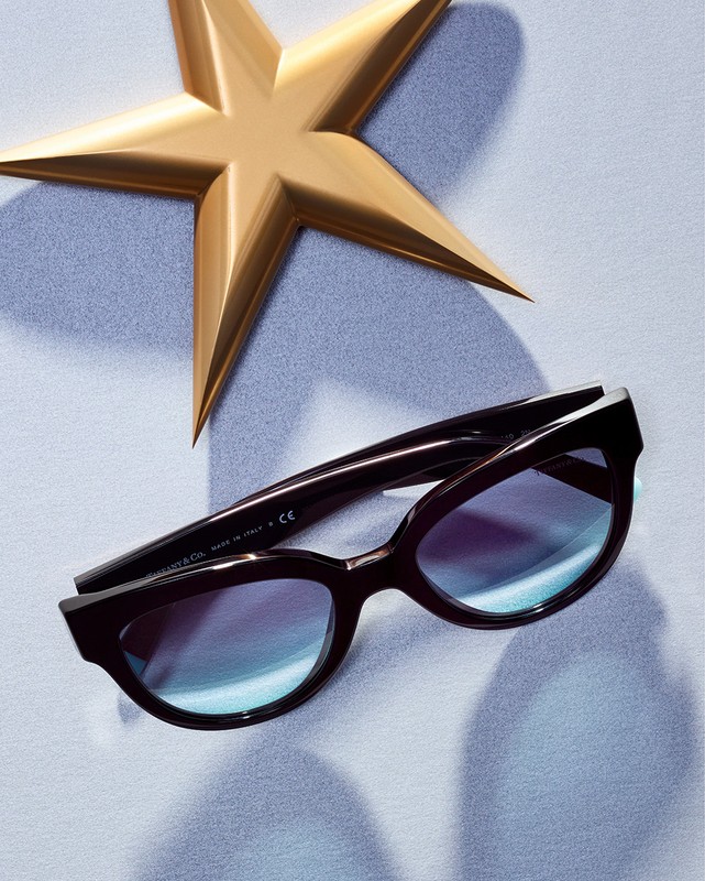 Studio shot of black sunglasses with blue gradient lenses next to a gold star-shaped object, all displayed on a light blue background.