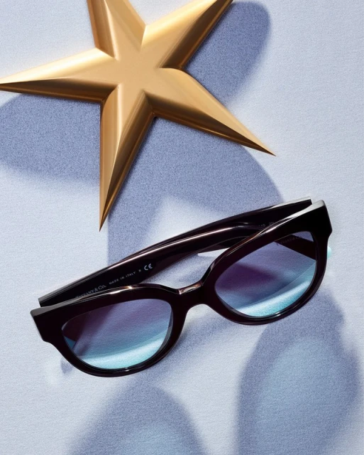 Studio shot of black sunglasses with blue gradient lenses next to a gold star-shaped object, all displayed on a light blue background.