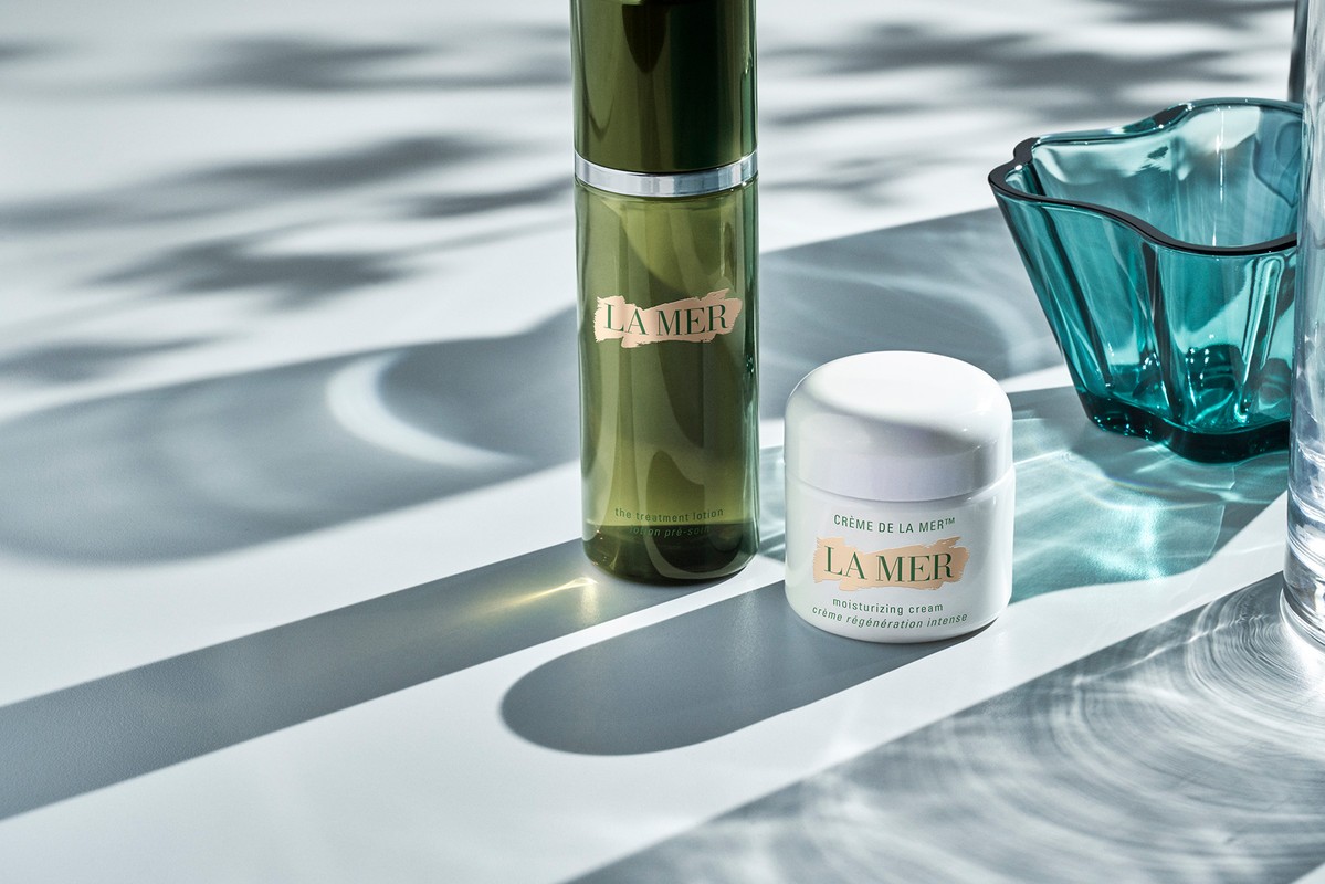 La Mer skincare products: a green bottle and white jar on a white surface with dappled light and shadows, and a blue decorative glass.