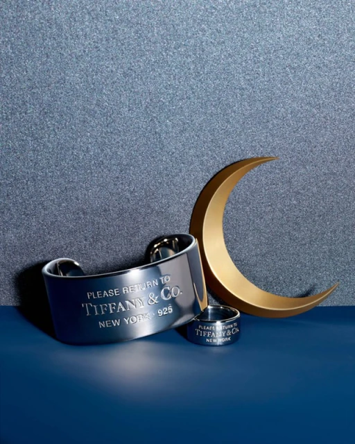 Close-up of Tiffany & Co. jewelry: a silver cuff bracelet and ring, with a gold crescent shape against a blue glitter background.