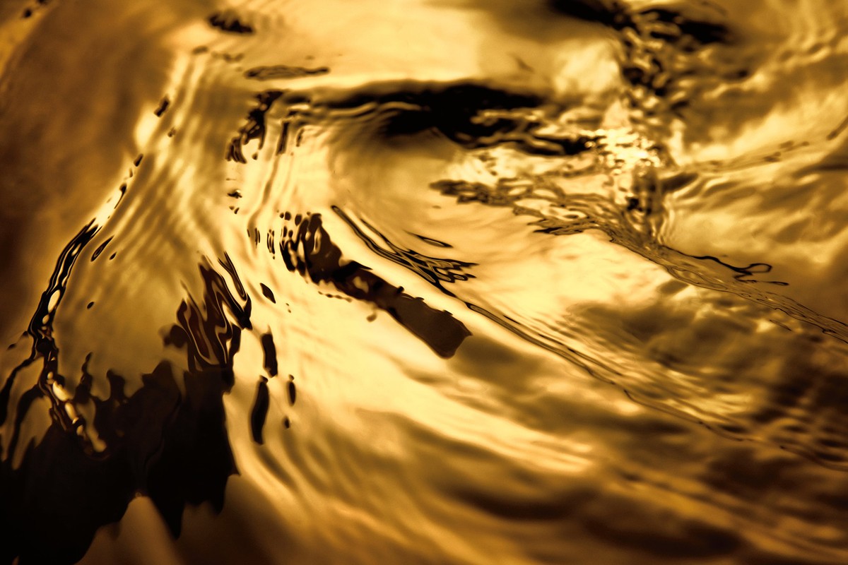 Abstract close-up of golden liquid surface with rippling texture and dark shadows, creating a flowing, metallic effect.