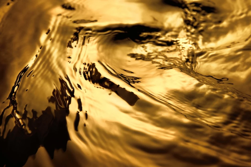 Abstract close-up of golden liquid surface with rippling texture and dark shadows, creating a flowing, metallic effect.