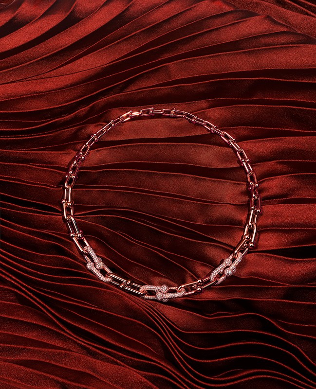 Close-up of a rose gold chain link necklace with diamond accents, displayed on a dark red pleated fabric background.