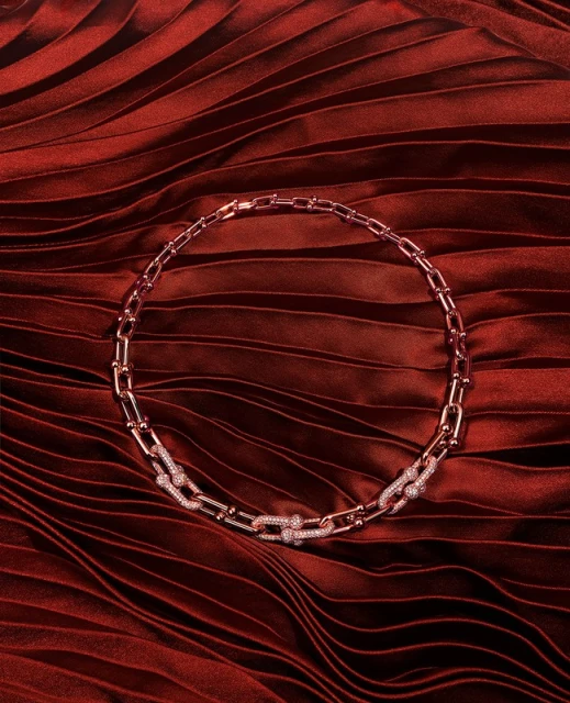 Close-up of a rose gold chain link necklace with diamond accents, displayed on a dark red pleated fabric background.