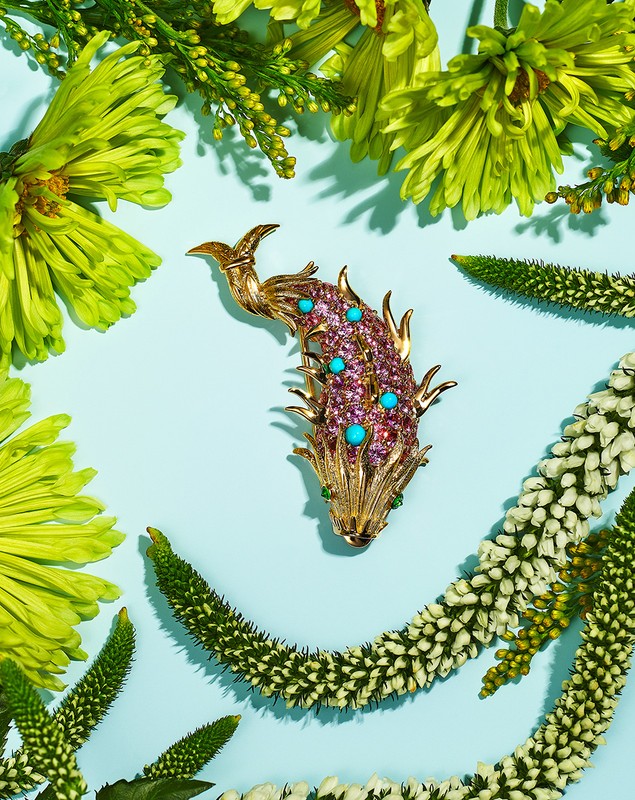 Ornate gold fish brooch with pink and turquoise gems surrounded by green and white flowers on a light blue background in a studio shot.