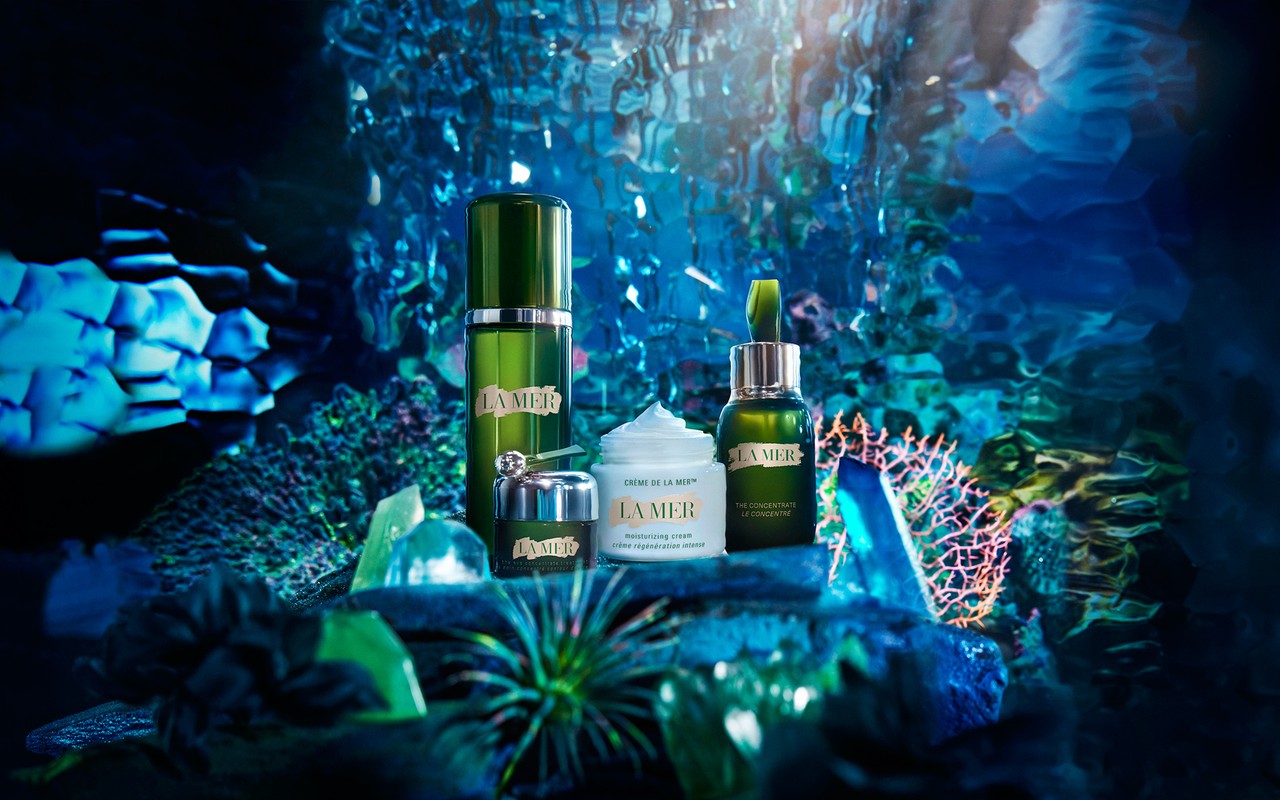 La Mer skincare product line displayed on a blue textured surface with a matching backdrop, featuring creams and serums in green packaging.