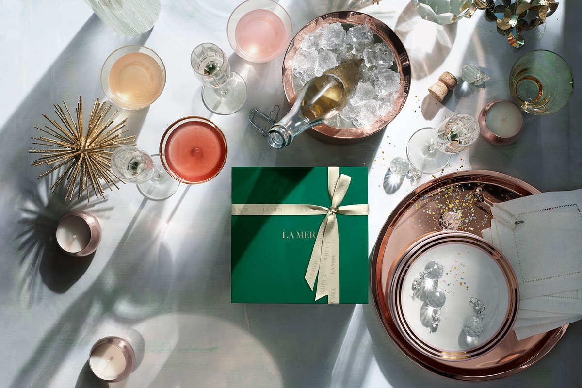 Festive still life of a La Mer gift box, glassware with cocktails, champagne bottle in an ice bucket, and candles on a white surface.