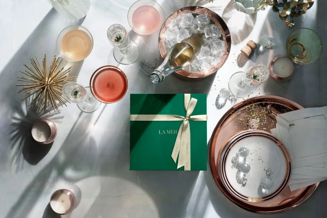 Festive still life of a La Mer gift box, glassware with cocktails, champagne bottle in an ice bucket, and candles on a white surface.