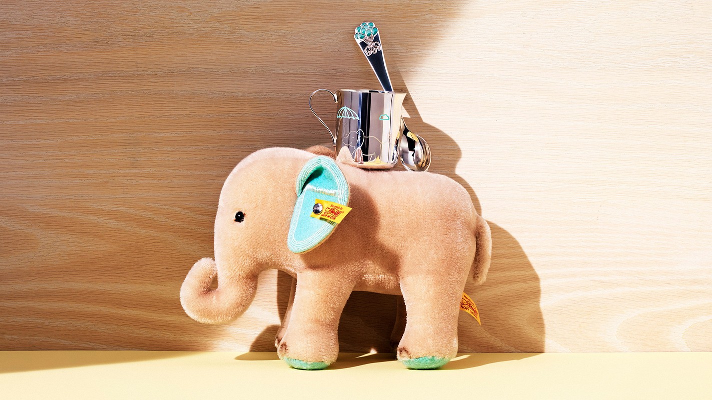 Still life of a plush beige elephant toy with blue details and silver mug, spoon, and teaspoon, set against a two-tone wooden backdrop.