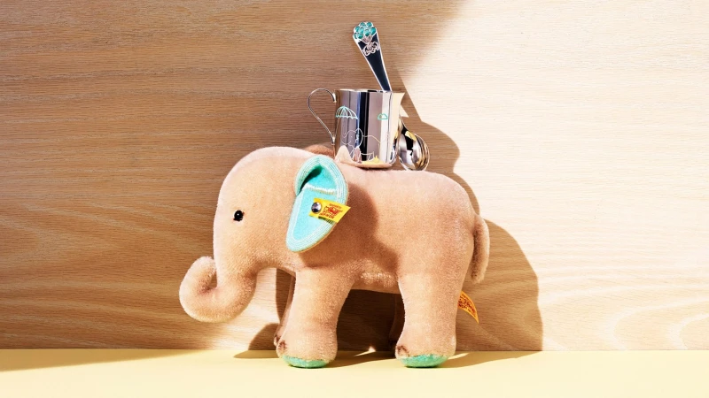 Still life of a plush beige elephant toy with blue details and silver mug, spoon, and teaspoon, set against a two-tone wooden backdrop.