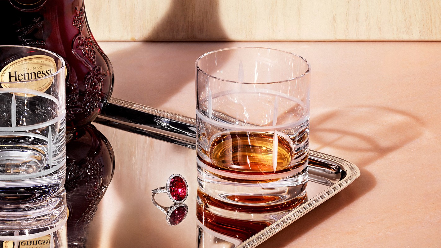 Crystal glass filled with cognac beside a luxury ring on a silver tray, and a dark bottle of Hennessy in a sophisticated setting.