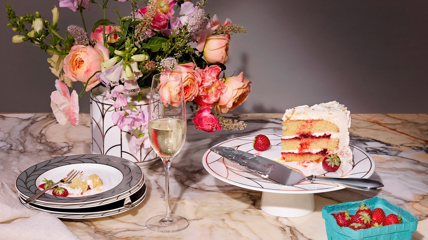 Celebratory still life: champagne flute, cake slice, and strawberries on marble surface, with a flower arrangement in the background.