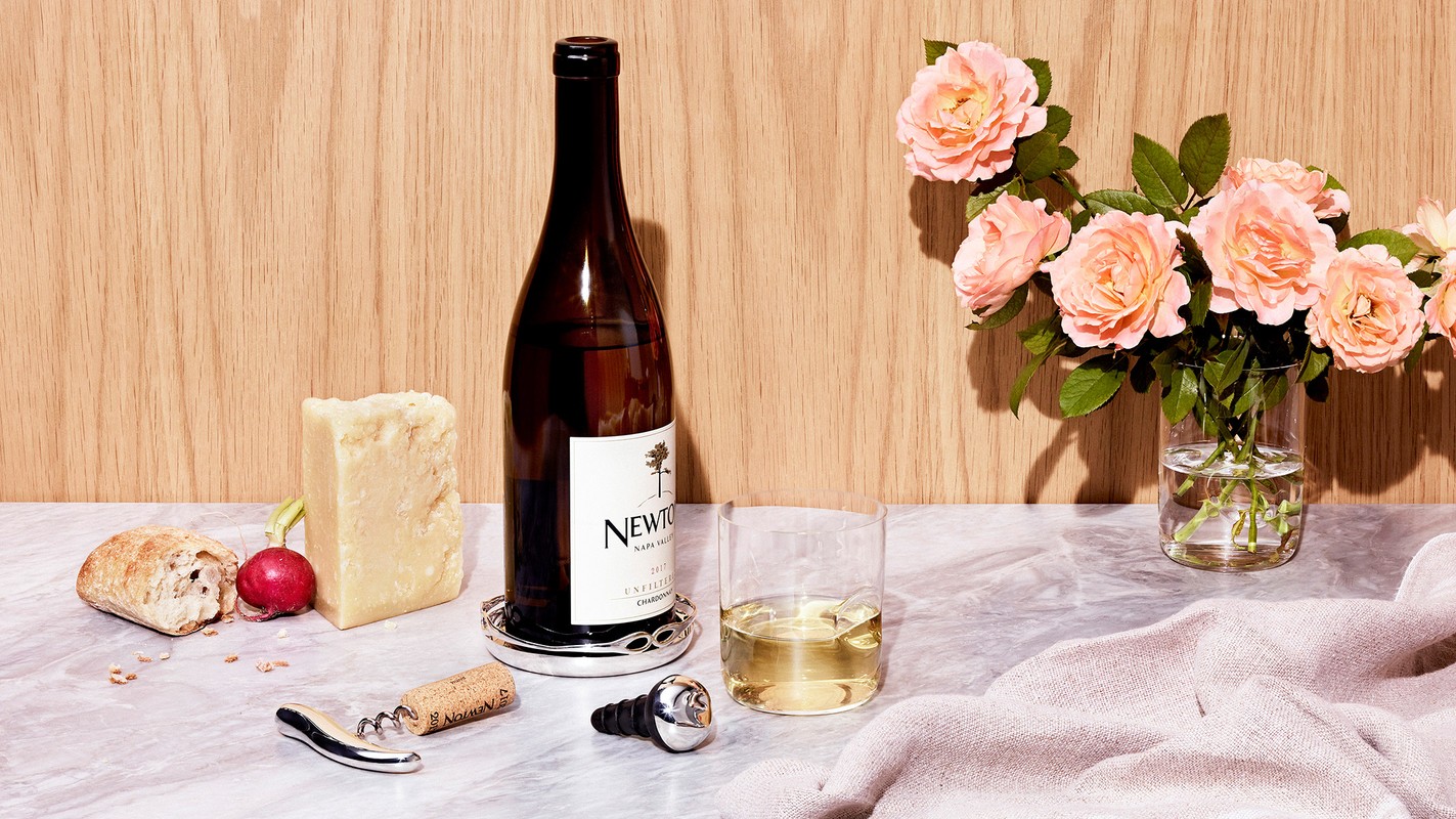 Wine bottle, glass of wine, cheese, bread, and flowers on a marble surface against a wood panel background. Still life.