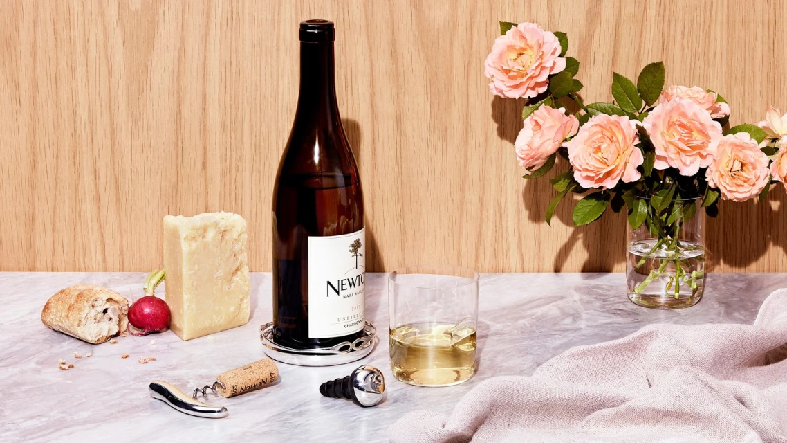 Wine bottle, glass of wine, cheese, bread, and flowers on a marble surface against a wood panel background. Still life.
