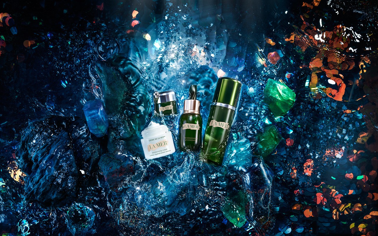 La Mer skincare products are displayed against a textured blue background with scattered gems, creating a luxurious, eye-catching still life.