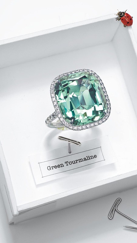Close-up of a green tourmaline ring with diamond halo displayed on a white surface with paper clip and a ladybug in the corner.