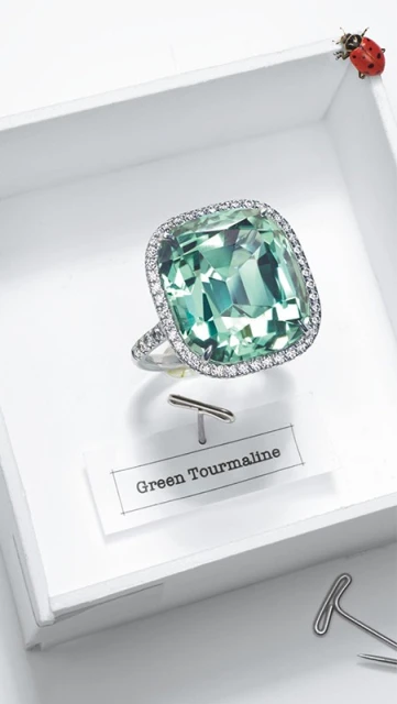 Close-up of a green tourmaline ring with diamond halo displayed on a white surface with paper clip and a ladybug in the corner.