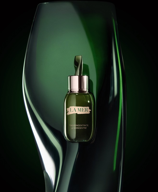 La Mer 'The Concentrate' serum bottle on a reflective green sculptural surface against a dark backdrop for a beauty product shot.