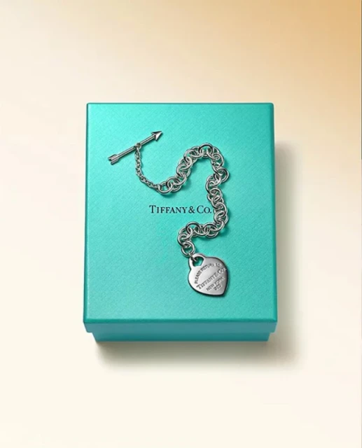 Silver chain bracelet with arrow and heart charm resting on a Tiffany & Co. turquoise gift box against a gradient background.