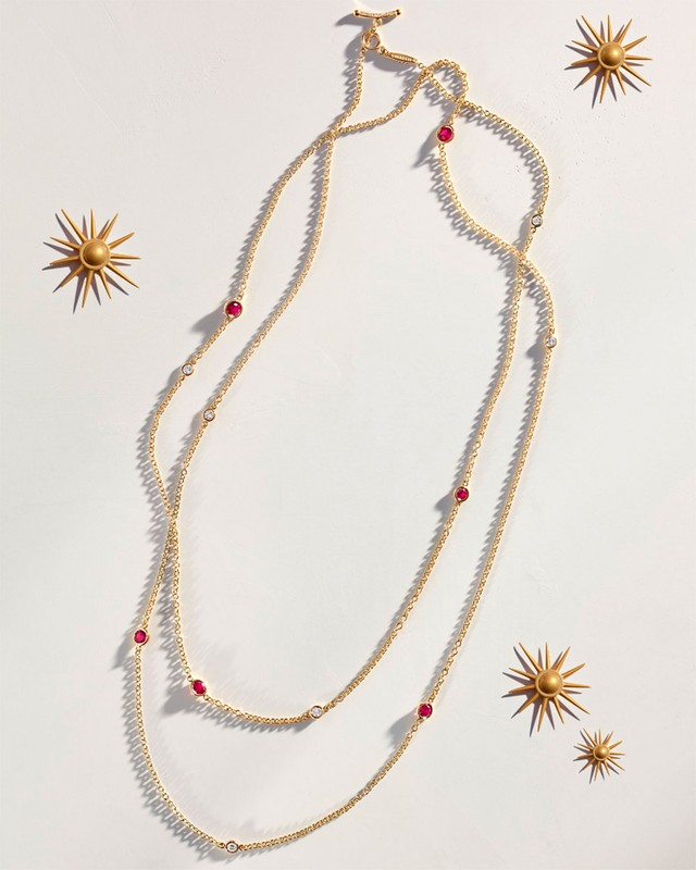 Gold necklace with ruby accents on a white background with decorative gold sunbursts, product photography for jewelry design.
