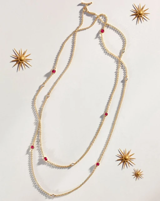 Gold necklace with ruby accents on a white background with decorative gold sunbursts, product photography for jewelry design.