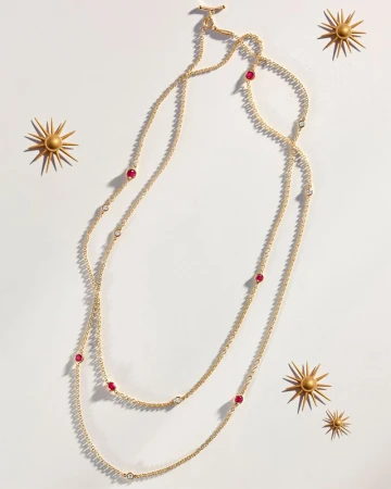 Gold necklace with ruby accents on a white background with decorative gold sunbursts, product photography for jewelry design.