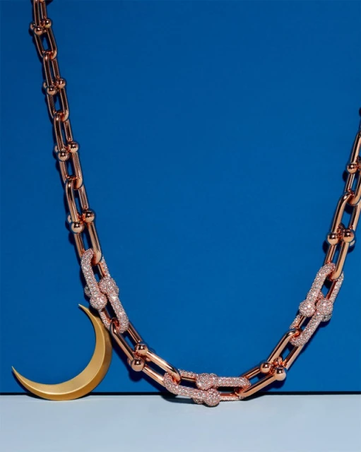Elegant rose gold chain-link necklace with sparkling accents, paired with a gold crescent moon pendant on a two-tone blue background.