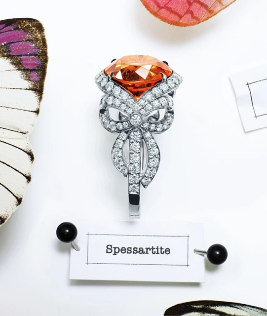 Statement ring with an orange stone and diamond embellishments, pinned to a white surface near butterfly wings and a card with the word "Spessartite."