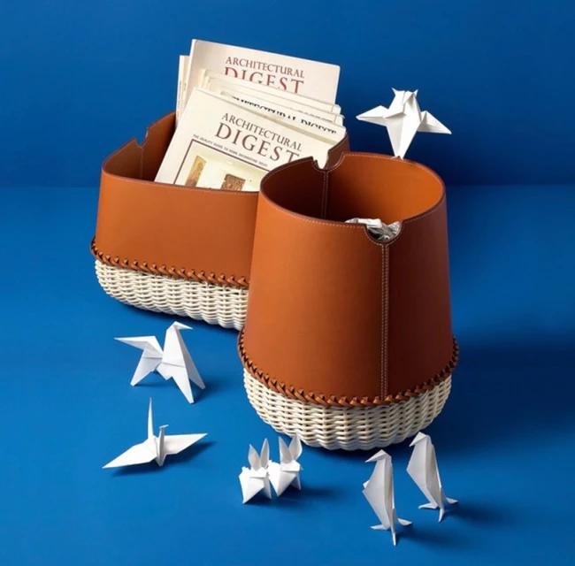 Two woven baskets with leather trim hold magazines, surrounded by origami paper sculptures against a solid blue backdrop. 