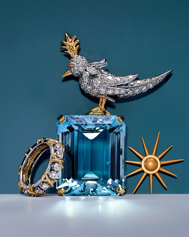Luxury jewelry still life: A jeweled bird figurine sits on a large aquamarine, with a diamond ring and a sunburst accent piece.