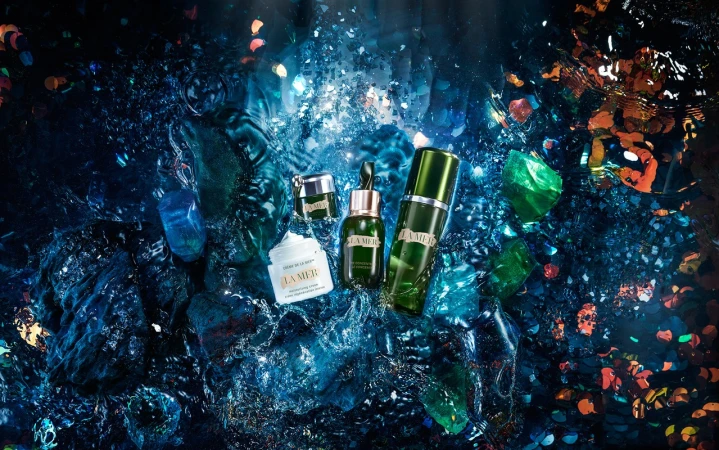 La Mer skincare bottles and jars resting amongst glittering blue and green crystalline water and floating confetti.