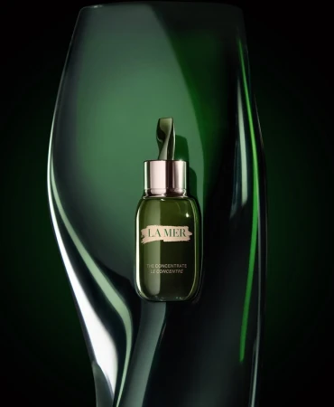Green glass skincare bottle with silver cap positioned against a dark glossy curved green glass background.