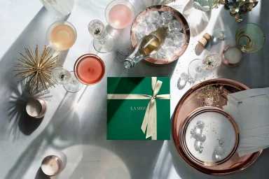 Top-down view of a La Mer green gift box surrounded by champagne glasses, ice bucket, and copper trays on a marble surface.