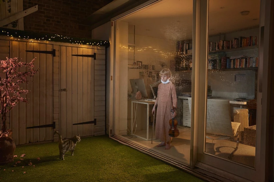 Indoor shot of a child holding a violin standing behind glass doors, with a cat sitting on grass outside.